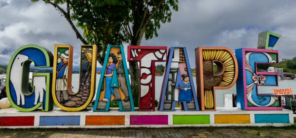 Colorful GUATAPE town sign with decorative local scenes, set beside the reservoir under a cloudy sky