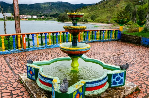 Colorful fountain with intricate detailing, surrounded by vibrant railings and green hills, overlooking a tranquil lake in Guatapé.