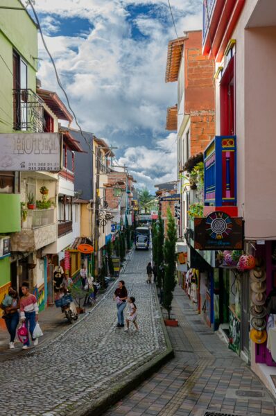 Colorful buildings line the cobblestone street in Guatapé, Antioquia, with people strolling and vibrant shops on either side.