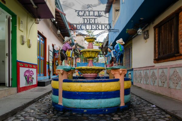 Colorful alley in Guatapé with a decorated fountain, painted façades, and a hanging Calle del Recuerdo sign