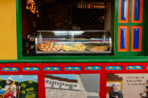 Colorful storefront in Guatapé, Colombia, featuring a display of pizzas and pastries, with vibrant murals and decorations.