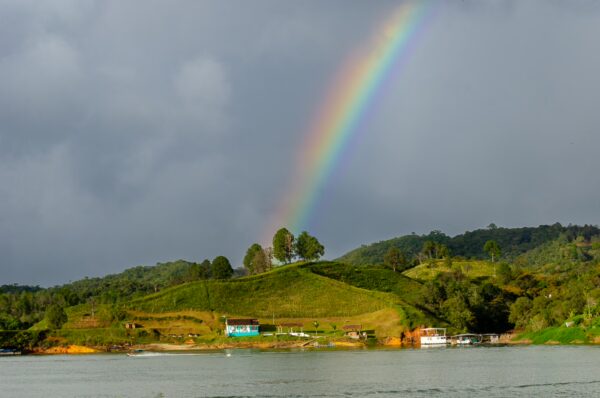 A vibrant rainbow arches over lush green hills with scattered trees near the water's edge in Guatapé, Antioquia, Colombia.