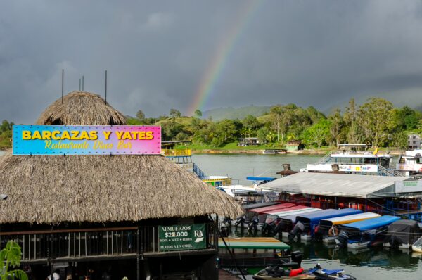 Colorful boats lined up at the marina in Guatapé, Colombia, with a vibrant rainbow arching over lush green hills.