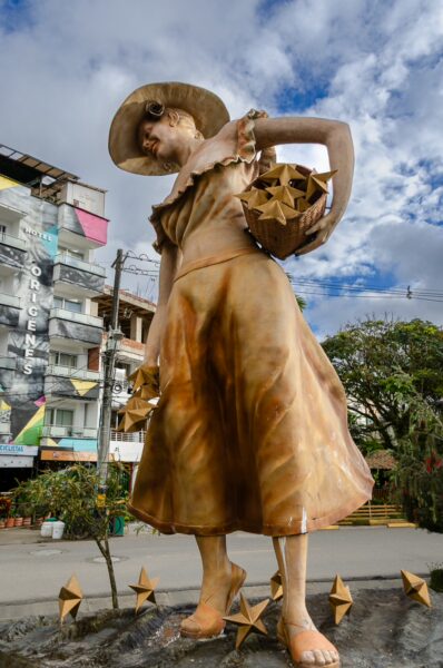 Large gold-colored sculpture of a woman holding a basket of stars in Guatapé, with a hotel building and cloudy sky behind it