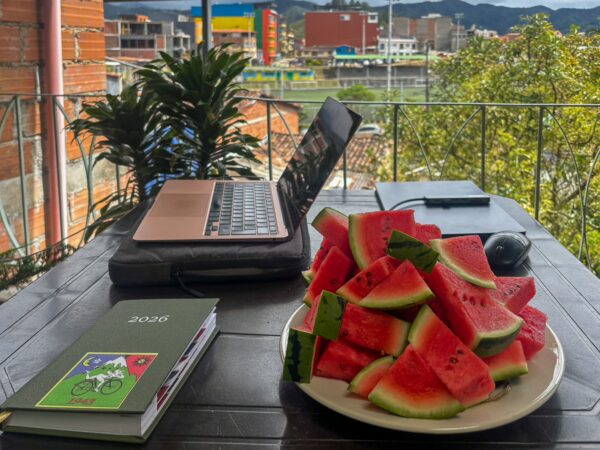Balcony table with a laptop, notebook with hofman cyclist sticker, mouse, and plate of watermelon slices overlooking Guatapé