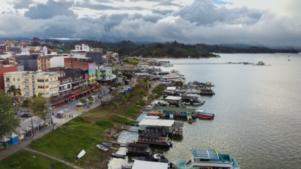 Colorful buildings and boats line the waterfront in Guatapé, Antioquia, with lush green hills and a cloudy sky in the background.