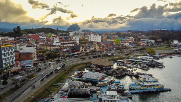 Vibrant Guatapé town features colorful buildings lining the waterfront, with boats anchored along the shore under a cloudy sky.
