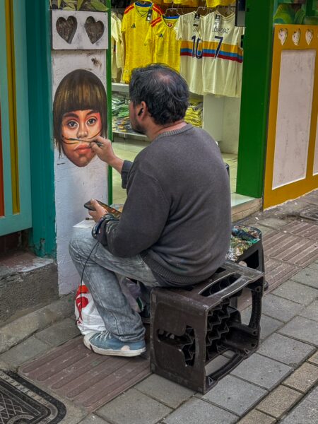 A man paints a colorful mural of a girl on a wall in Guatapé, while vibrant shirts hang in a shop nearby.