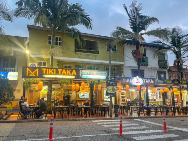 Colorful buildings with tropical palm trees line the streets of Guatapé, featuring outdoor dining at Tiki Taka under warm lights.