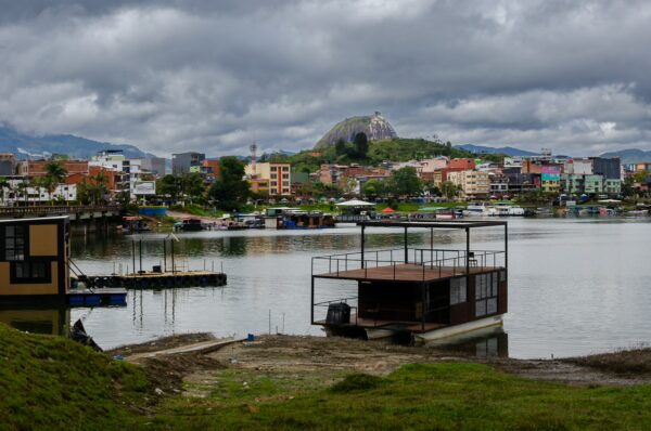 Colorful buildings line the shore of Guatapé, reflecting in the calm waters under a cloudy sky, with a distant hill crowned by an iconic rock.