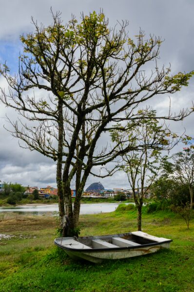 A small boat rests on grass by the reservoir, with Guatapé and El Peñón in the distance under a cloudy sky