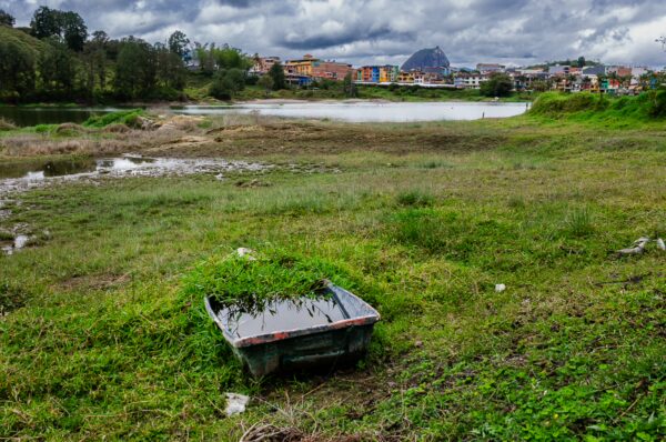 Lush green grass surrounds a neglected boat by the waterside, with colorful buildings and hills in the background near Guatapé, Colombia.