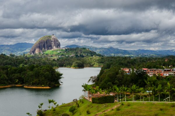 Cloudy skies loom over Guatapé, with a striking rock formation rising above lush green hills and a serene lake.