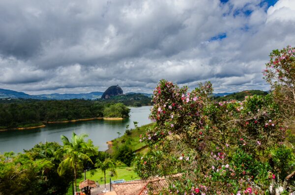 Lush greenery and vibrant flowers frame the tranquil lake near Guatapé, with rolling hills and blue skies above.