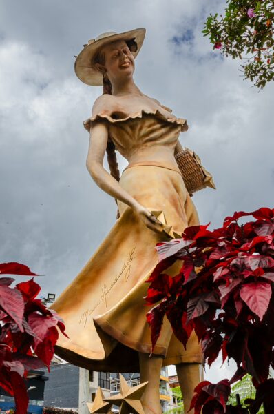 Dramatic statue of a woman in a flowing dress, surrounded by vibrant red plants, set against a cloudy sky in Guatapé, Colombia.