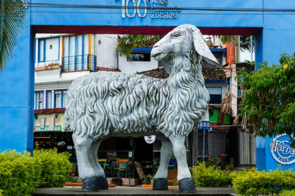 A large, whimsical sheep sculpture stands in front of colorful buildings in Guatapé, Colombia, surrounded by vibrant greenery.