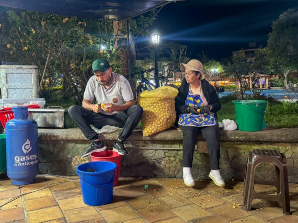 Two vendors sit by buckets in Guatapé’s main square at night, with a sack of potatoes and lit trees behind them