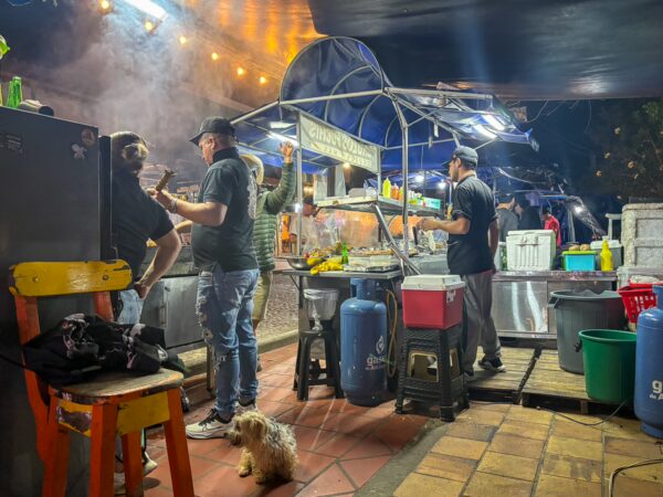 A vibrant street food stall in Guatapé, with chefs preparing meals under warm lights, while a small dog sits nearby.