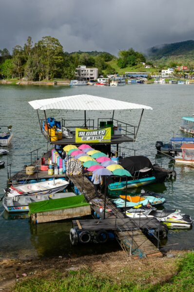 Colorful umbrellas shade a vibrant dock in Guatapé, Colombia, with boats moored on the serene water, surrounded by lush greenery.