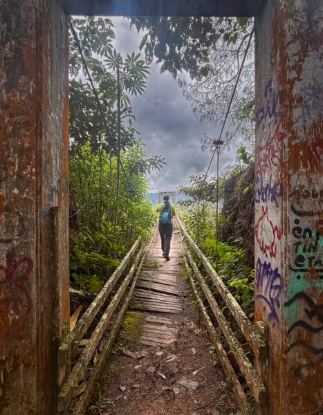A person walks along a rustic wooden bridge surrounded by lush greenery and colorful graffiti in Guatapé, Colombia.