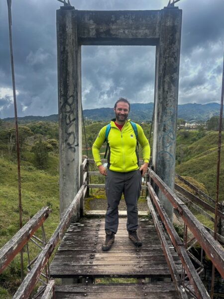 A man in a bright green jacket stands on a weathered suspension bridge surrounded by lush greenery in Guatapé, Colombia.