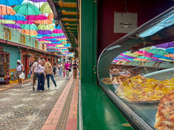 Colorful umbrellas hang above a vibrant street in Guatapé, Colombia, with people walking past a bakery displaying fresh pastries.