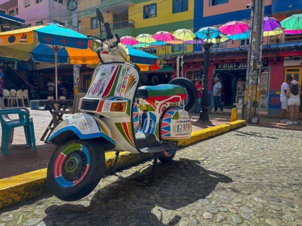 Vibrant umbrellas hang over colorful buildings in Guatapé, with a decorated scooter parked on cobblestone streets.