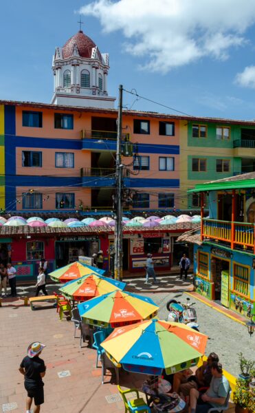 Vibrant buildings line the Plaza de los Zócalos in Guatapé, Colombia, featuring colorful umbrellas and a historic dome in the background.