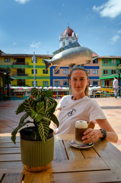 Woman sitting at an outdoor café in Plazoleta de Los Zócalos, with a fish statue and colorful Guatapé buildings behind her
