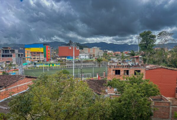 Colorful buildings surround a green sports field in Guatapé, Antioquia, under a dramatic cloudy sky, with mountains in the background.