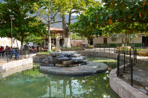 A shady village square in Jordán with a circular fountain, stone statue, benches, and people gathered under large trees
