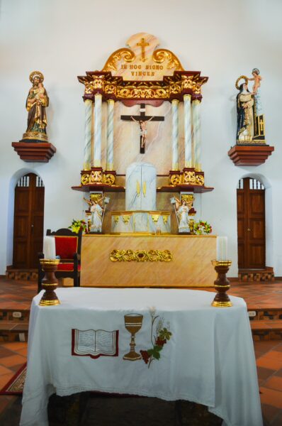 Altar of a church in Santander, Colombia, featuring ornate golden accents, statues, candles, and a decorative altar cloth.