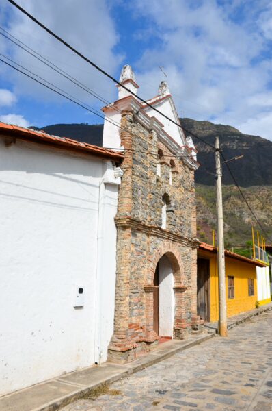 Historic stone structure beside vibrant yellow building in Santander, Colombia, framed by a cloudy sky and mountainous backdrop.