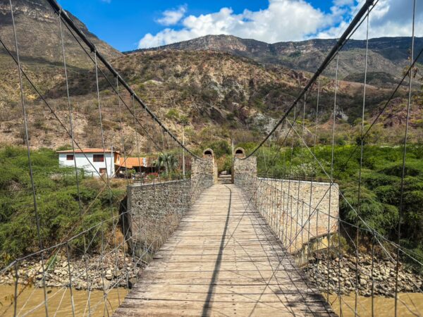 Suspension bridge in Santander, Colombia, spanning a river with lush greenery and mountainous scenery in the background.