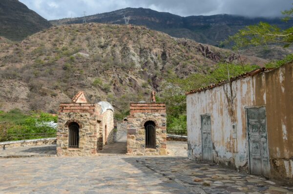 Jordan, Santander, Colombia: Stone structures with arched openings against a mountainous backdrop and cloudy sky. Lush greenery visible.