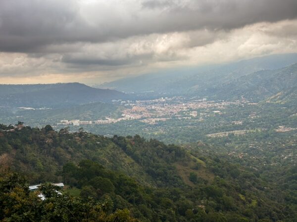 Wide view from Mesa Los Santos over green hills, a valley town, and distant mountains under heavy clouds