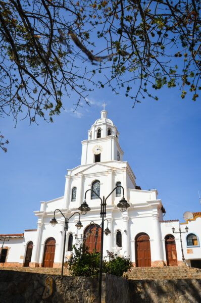 White church with clock tower in Los Santos square, framed by tree branches against a blue sky