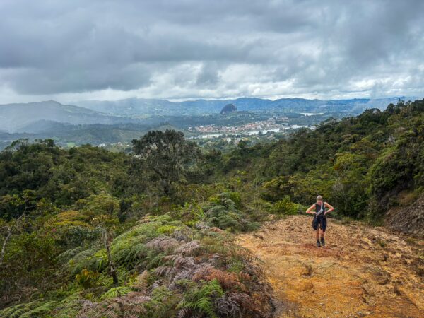 Mirador San José, Guatapé, Antioquia: A hiker on a rugged path overlooks lush green hills and a distant village under a cloudy sky.