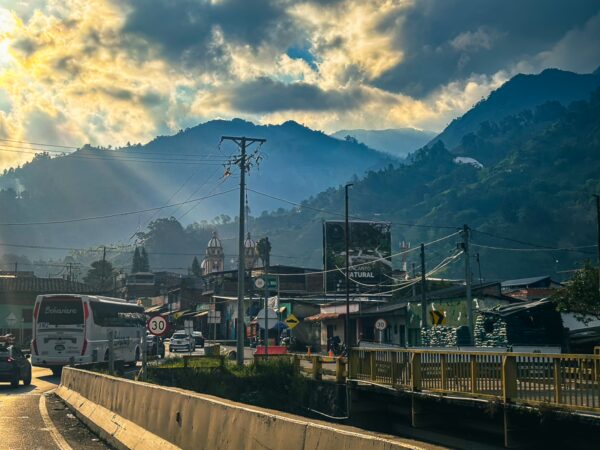 Cajamarca, Tolima features rolling mountains in the background with bustling streets and vehicles under a dramatic sky.