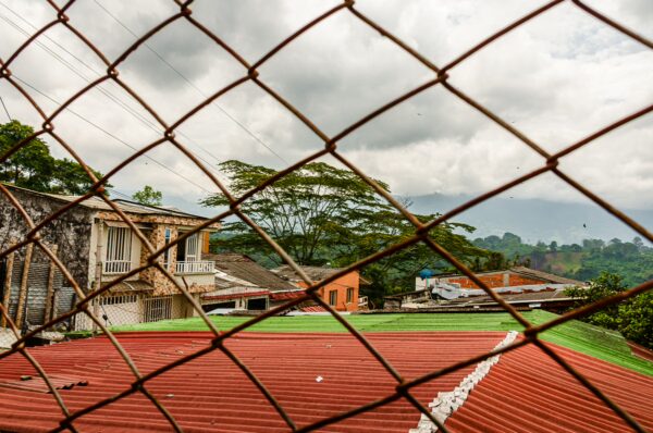 Rustic buildings with colorful roofs peek through a rusty fence, framed by lush green hills under a cloudy sky in Quindio, Colombia.