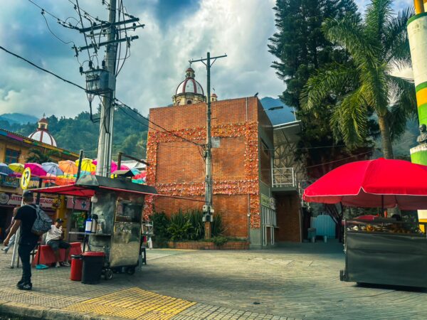 Vibrant street scene in Cajamarca, Tolima, Colombia, featuring a food cart, colorful umbrellas, and lush palm trees.