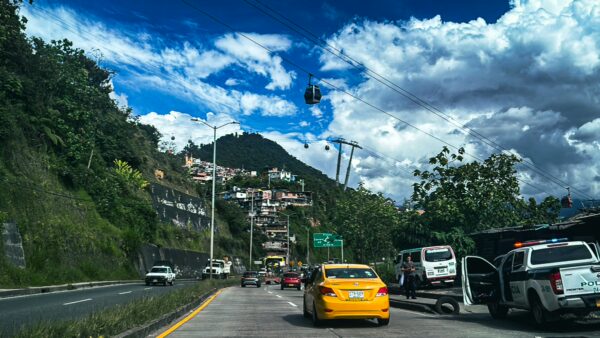 Bright yellow taxi on a road in Manizales, Colombia, with lush green hills and a cable car in the background under blue skies.