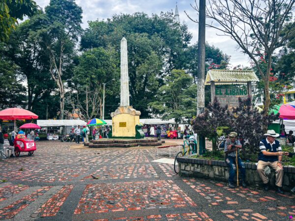 Vibrant Parque Principal in Líbano, Tolima, features a tall monument, local vendors, and lush greenery, creating a lively atmosphere.