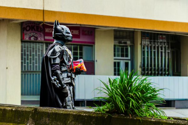 A figure in a Batman costume stands with snacks in Quindío, Colombia, against a backdrop of urban buildings and greenery.