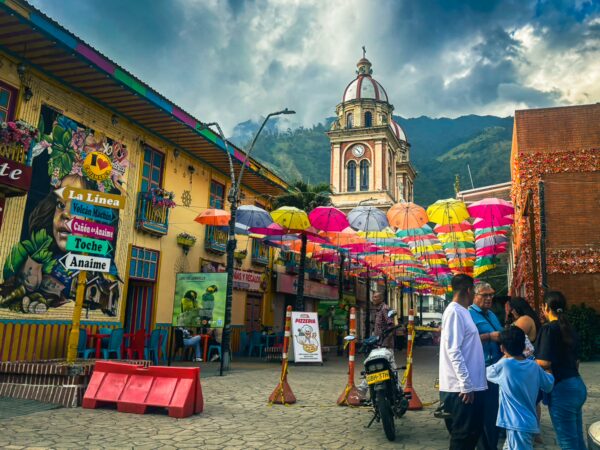 Colorful umbrellas hang above a vibrant street in Cajamarca, Tolima, Colombia, with a church tower visible against the mountains.