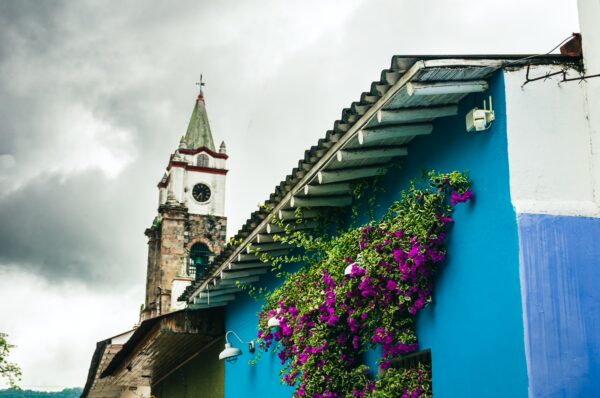 Colorful blue walls adorned with vibrant purple flowers and a church tower in Honda, Tolima, Colombia under a cloudy sky.