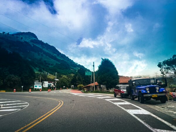 Winding road in Manizales, Caldas, Colombia, featuring lush green mountains, a blue jeep, and a scenic sky.