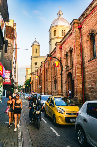 Colorful street scene in Pereira, Risaralda, featuring brick buildings, a yellow taxi, and people walking along the busy road.