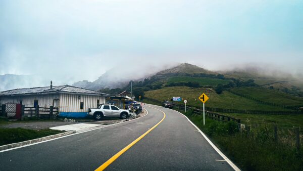 Curving road leads to lush green hills shrouded in mist near Nevado del Ruiz, Tolima, Colombia. Quiet rural scene with a few buildings.