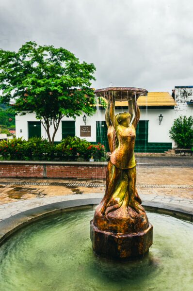 Golden fountain with a female figure in José León Armero Park, surrounded by greenery and colonial-style buildings in Honda, Tolima.